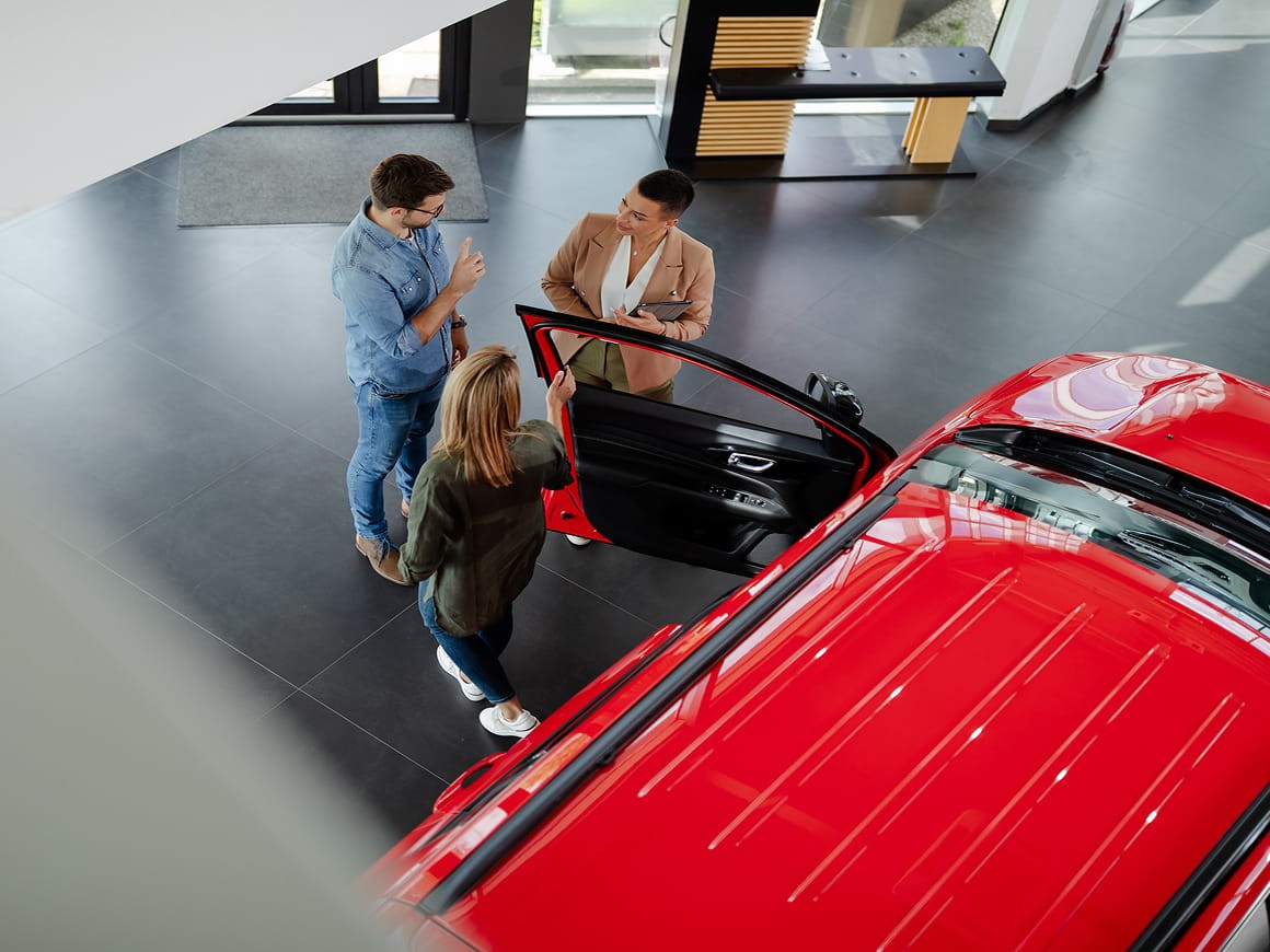 People discussing a vehicle beside a red car in a showroom while exploring options for salary packaging a car through a novated lease